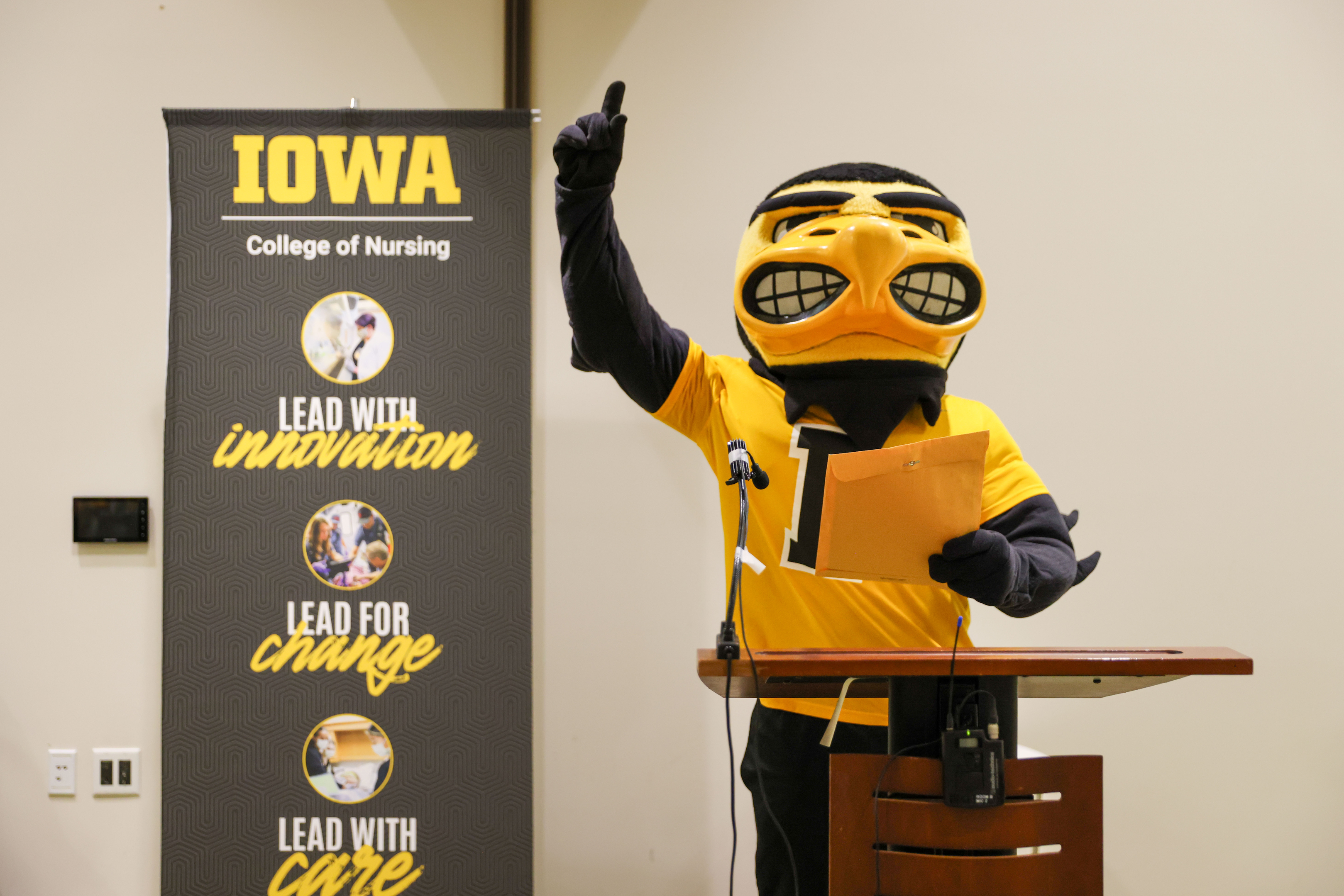 Herky stands at lecturn with finger in the air. College of Nursing banner is behind him. 